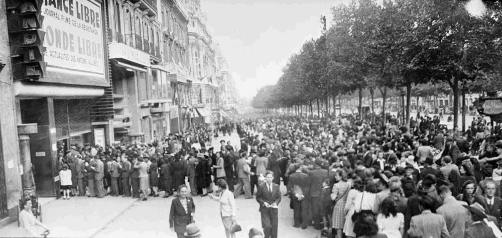 La foule déambule sur les Champs-Elysées pendant que des spectateurs attendent pour assiter à une séance de cinéma présentant des actualités de la France Libre et des Alliés, septembre 1944